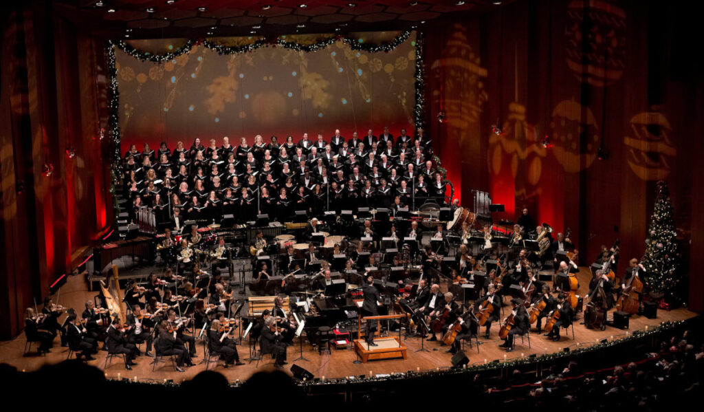 A symphony orchestra and choir are gathered on a holiday decorated stage