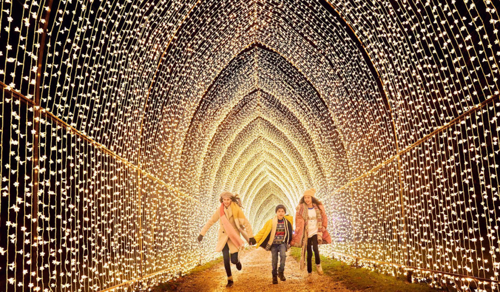 Three kids run through an arched tunnel of white holiday lights