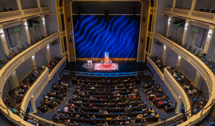 An auditorium filled with people watching two guests have a conversation on stage
