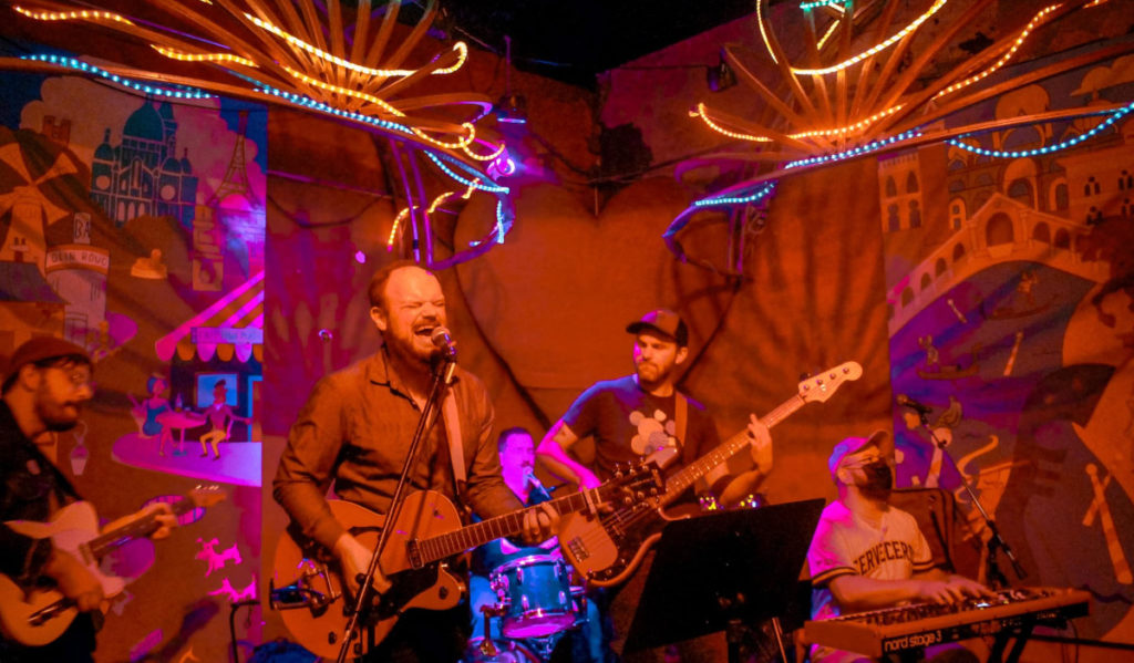 A band performs on a corner stage in orange and purple lights