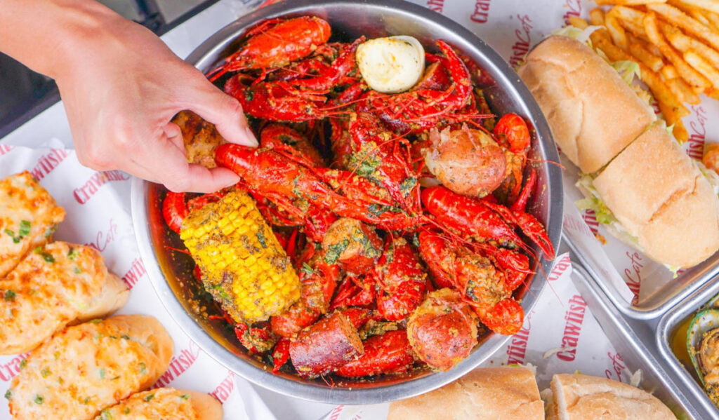 A hand reaches for a crawfish from a bowl, with other sandwich and fried offerings