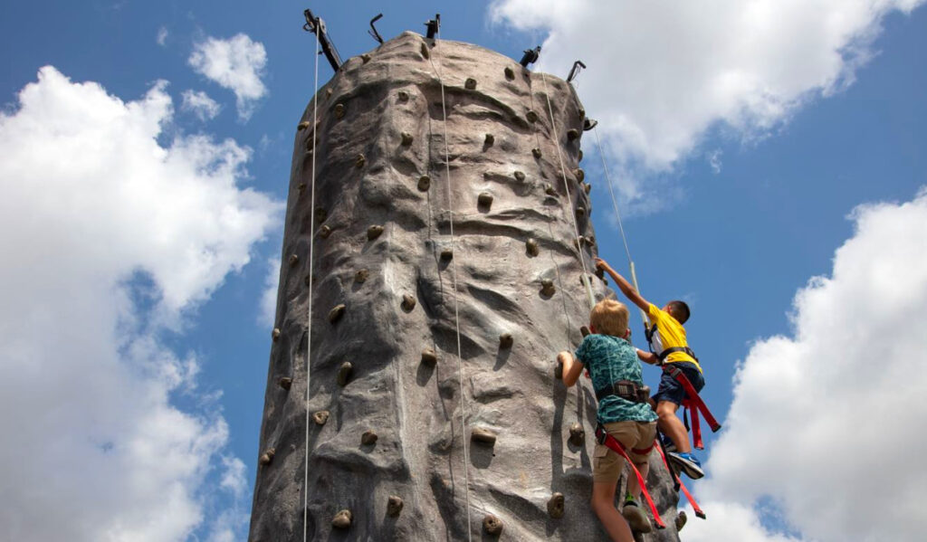 An angled shot upwards featuring two kids climbing a cylindrical rock wall with clouds in the background