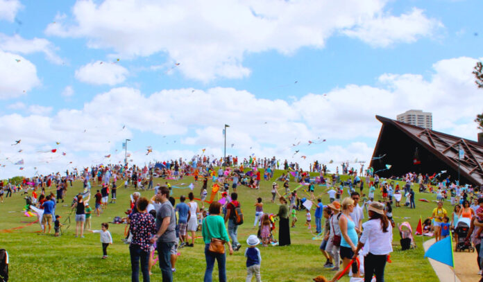A wide shot of families and children on the Hermann Park Hill with dozens of kits in the air