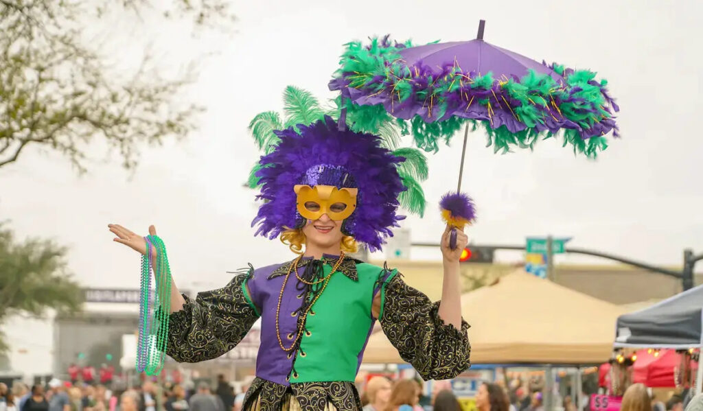 A stilt walker in green and purple Mardi Gras costume holding an umbrella