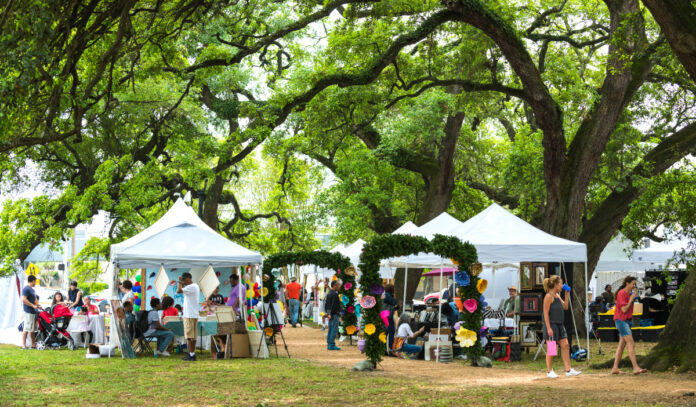 A market features stalls under a leafy canopy of tall oak trees