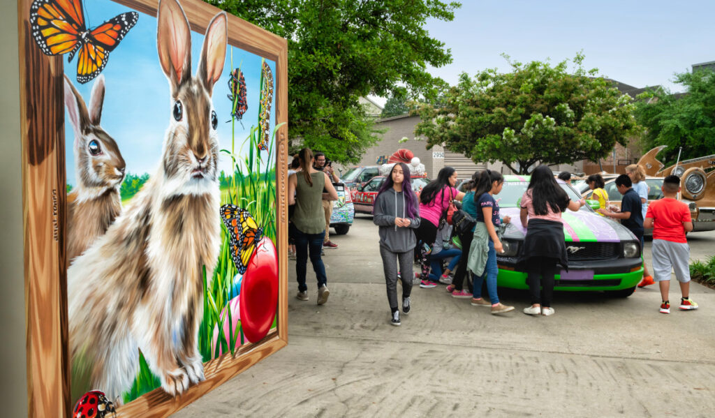 A teenager passes by a large mural of rabbits while kids write with chalk on a car