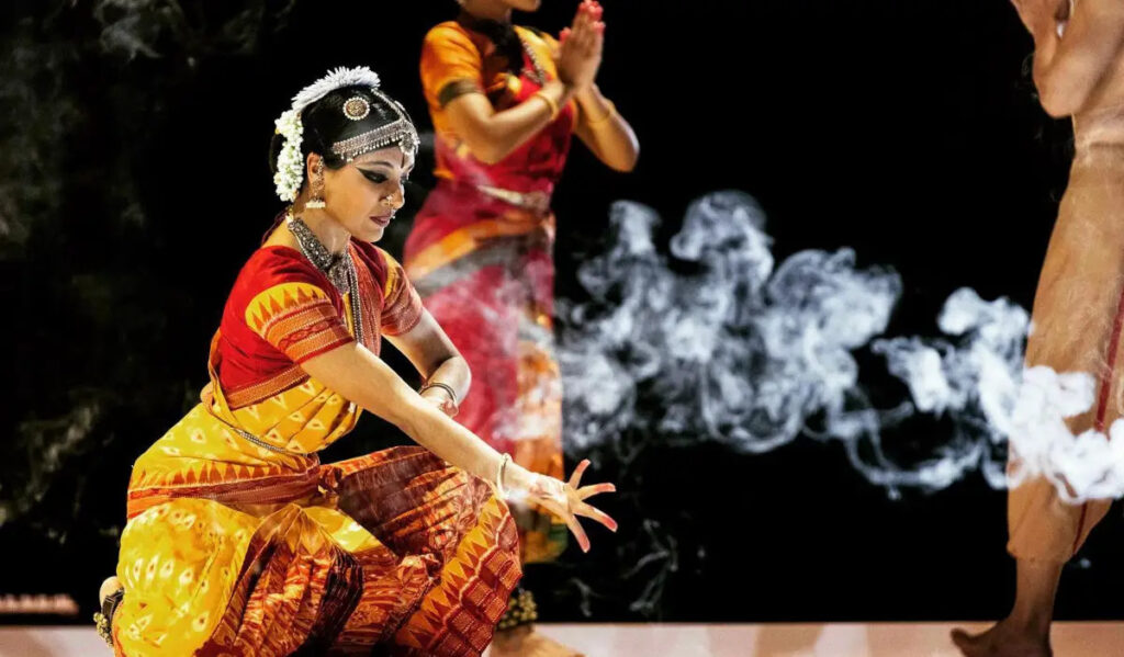 A performer in Bharatanatyam dress kneels near a cloud of smoke