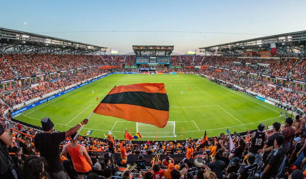 A supporter waves an orange and black flag in the foreground of a soccer stadium