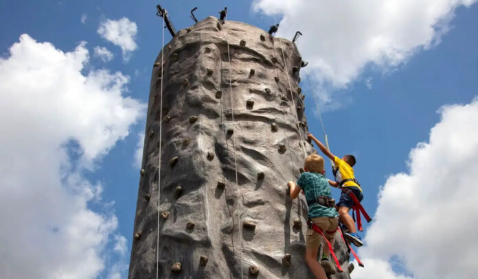 A low angle photo of two kids climbing a rock wall with clouds behind them