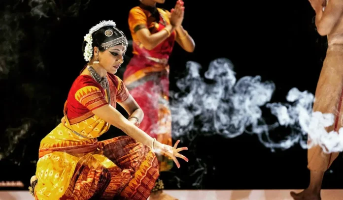 A performer in Bharatanatyam dress kneels near a plume of smoke