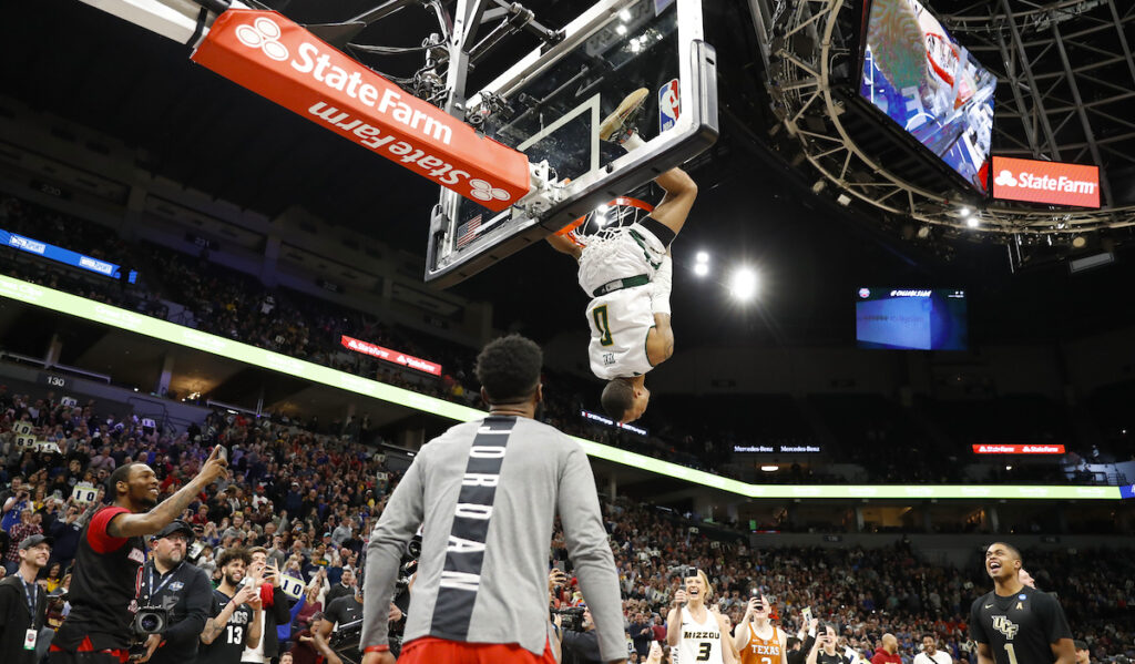 An athlete hangs upside down after dunking a basketball while people celebrate around him