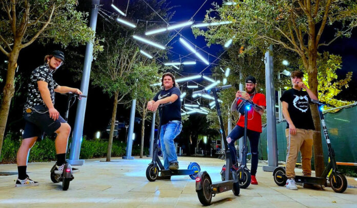 Four people pose with scooters under bars of light