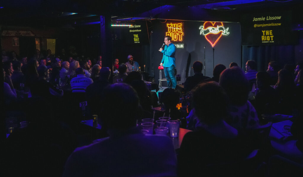 A stand-up comic performs in a dimly lit room in front of an audience