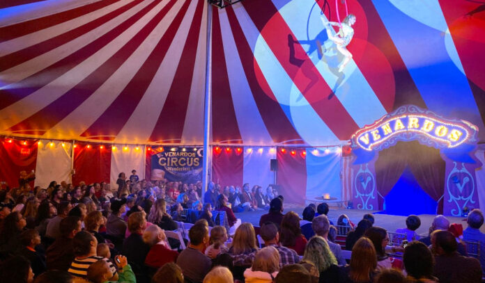 An aerial performer is suspended above a crowd inside a red and white striped tent
