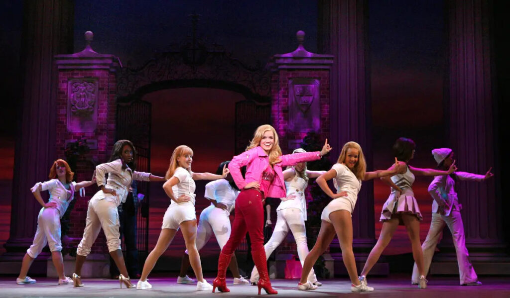 A troupe of dancers pose, looking to the camera over their shoulder with a pink-clad woman in the center