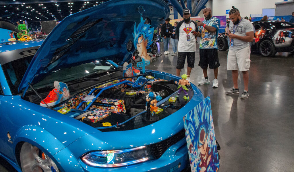 Three people discuss a custom car with its hood popped open and Dragon Ball Z art inside