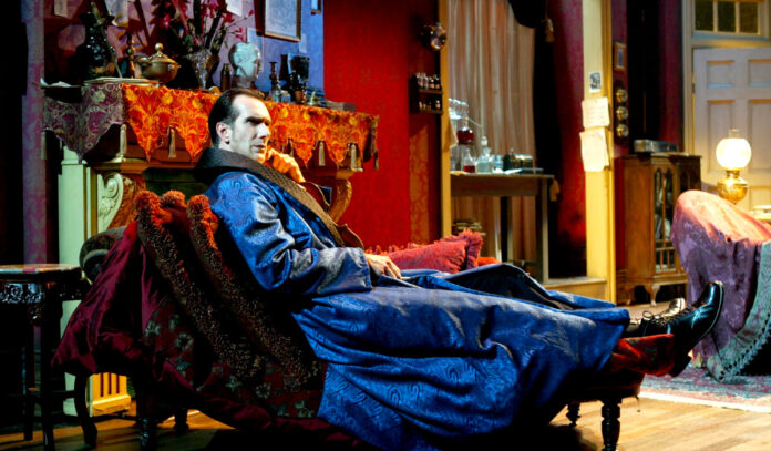 Todd Waite as Sherlock Rolmes, dressed in a robe and reclining in an armchair