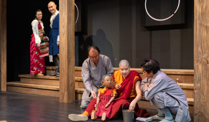 Two actors stand talking in the background while three others and a puppet sit in the foreground