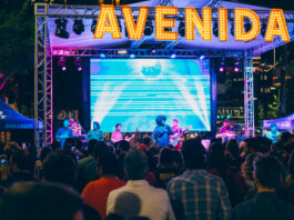 A brightly lit concert stage with performers under bulb lights that spell "Avenida"
