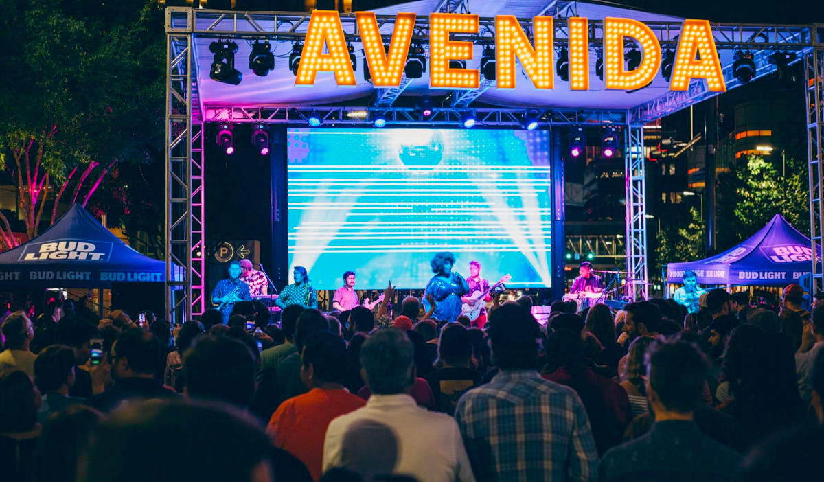 A brightly lit concert stage with performers under bulb lights that spell "Avenida"