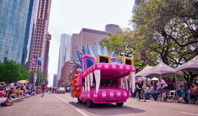 A pink Art Car with giant teeth and a large mouth rides through Downtown Houston streets