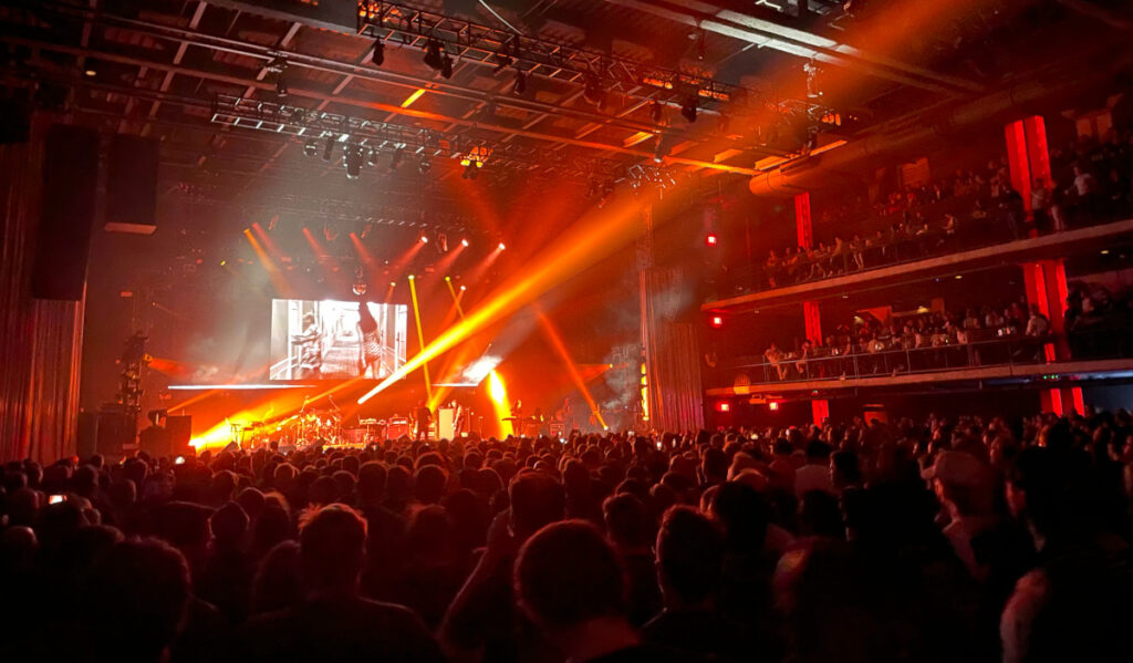 An arena concert with beams of orange light shining on the crowd as a band plays on stage