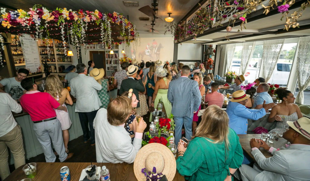 An interior of a bar with flower drapes and people in decorative hats watching the Kentucky Derby