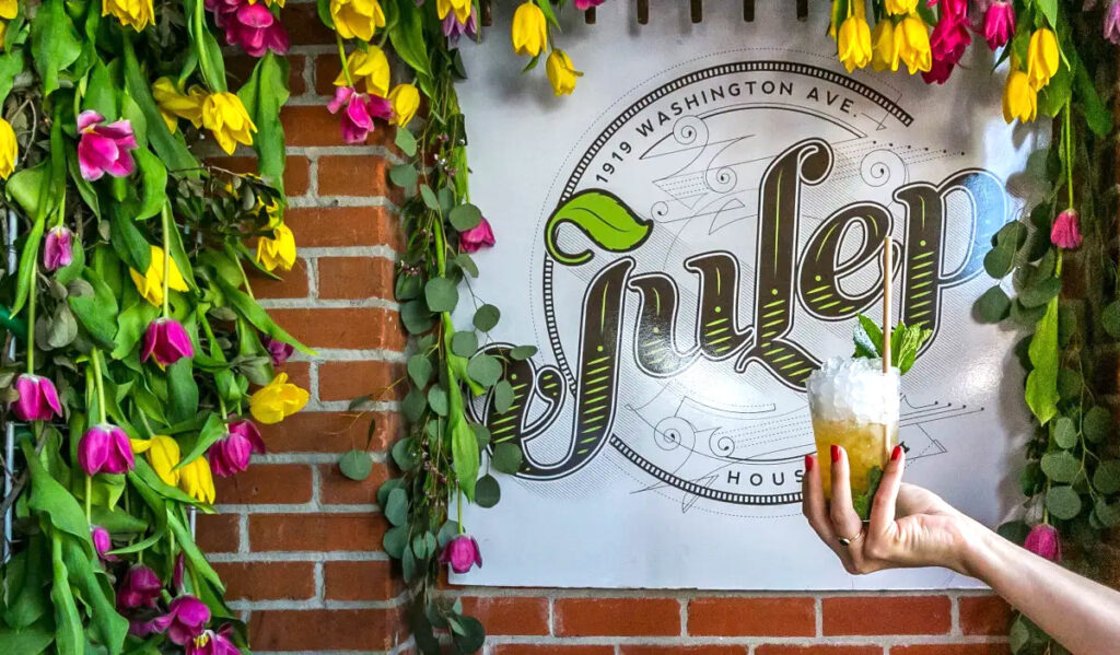 A hand holding a mint julep in front of the sign for the Julep bar