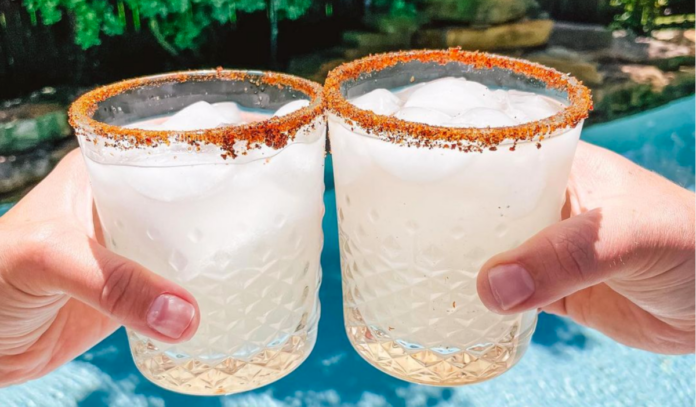 Two glasses of margaritas with tajin rims clink over a pool