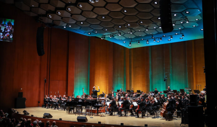 The Houston Symphony performs on stage with wall lighting in green, orange and blue behind them