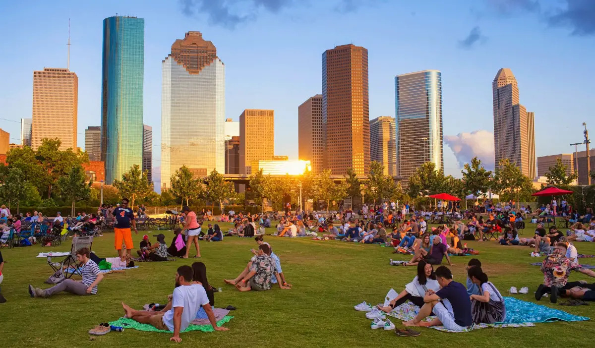 People sit on blankets on a lawn at sunset with a gleaming Downtown Houston skyline in the background