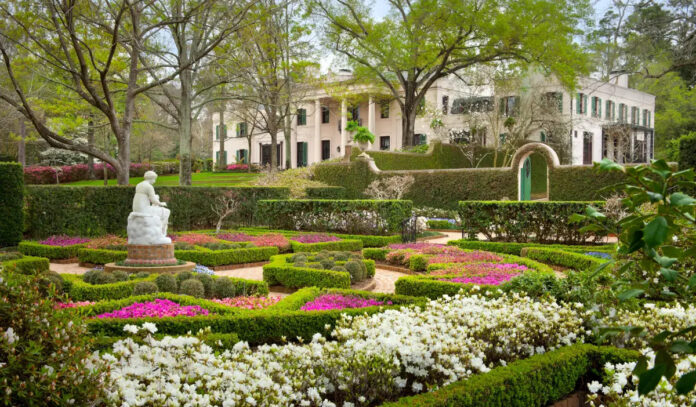 A highly manicured gardens of pink and white flowers with a plantation style house in the background