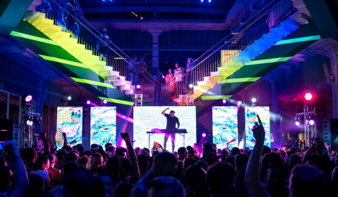 A DJ performs to a crowd with rainbow lights behind him