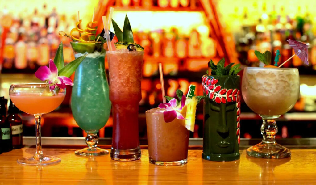 Six tiki drinks in different colors with fruit garnishes lined up on a bar