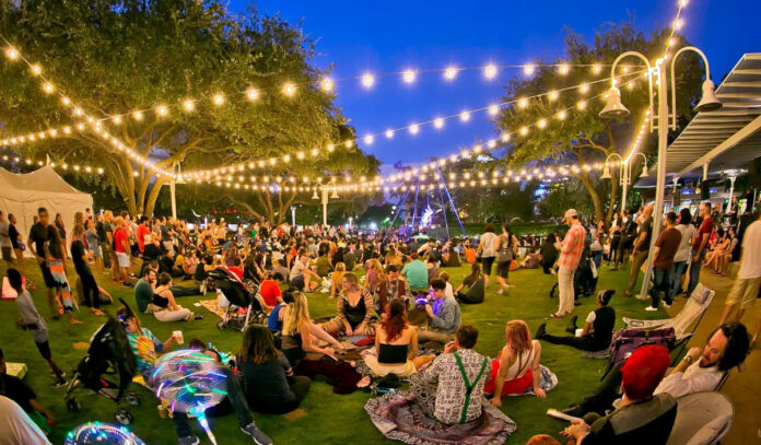 An evening scene with people in groups on a lawn and strings of lights overhead