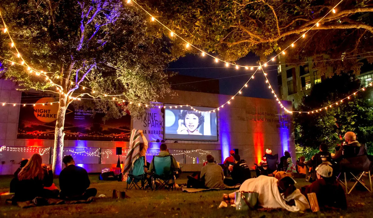 An outdoor movie screening on the side of a building as people watch from blankets and chairs under strings of lights