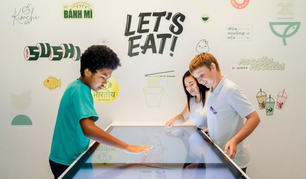 Three children interact with a touch screen in front of a wall with Asian cuisine words