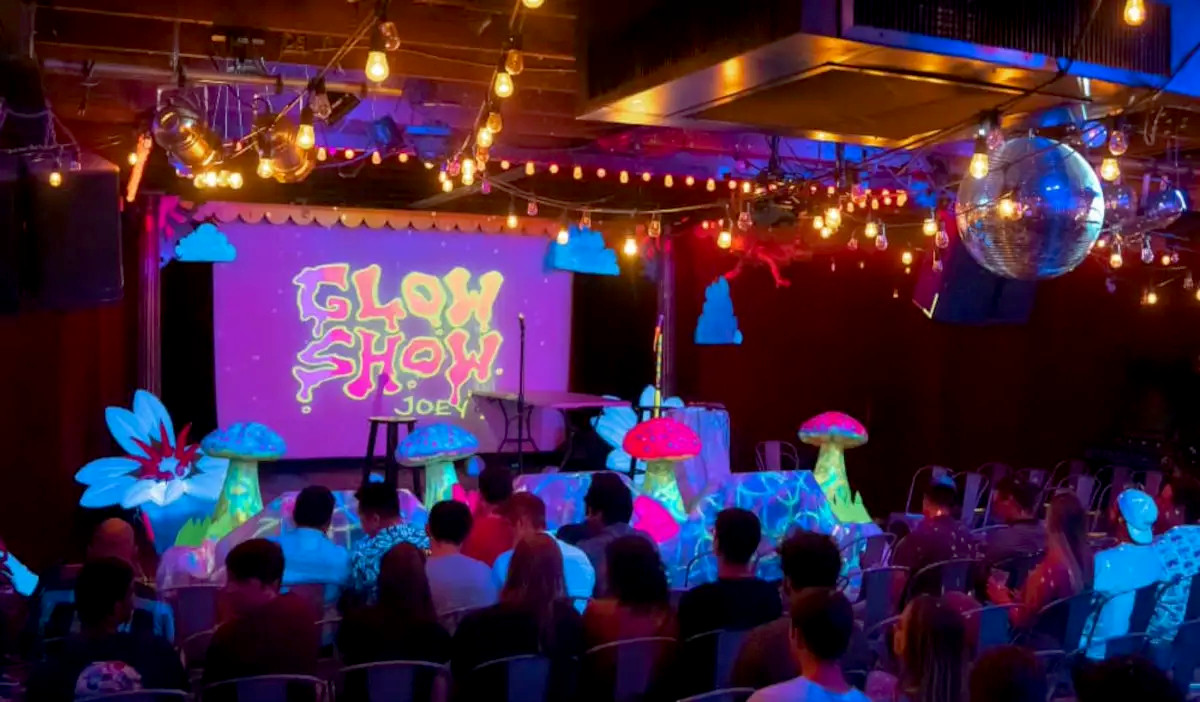 A crowd sat for a comedy show with UV light and glowing objects on stage