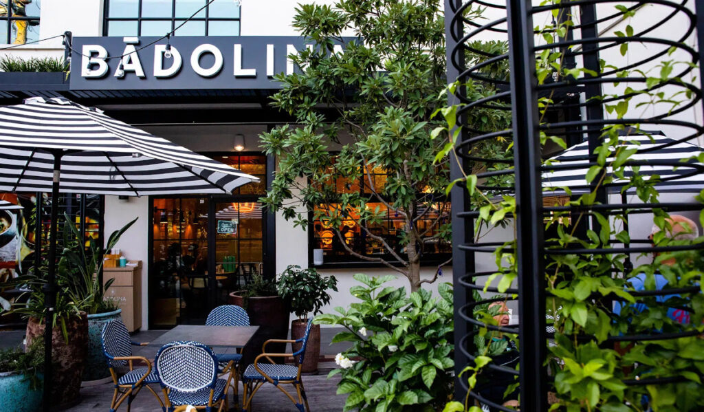 The exterior of Badolina Bakery with chairs, leafy plants and striped canopies