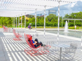 A person sits in a red chair within bands of sunlight near a fountain at a park