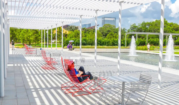 A person sits in a red chair within bands of sunlight near a fountain at a park