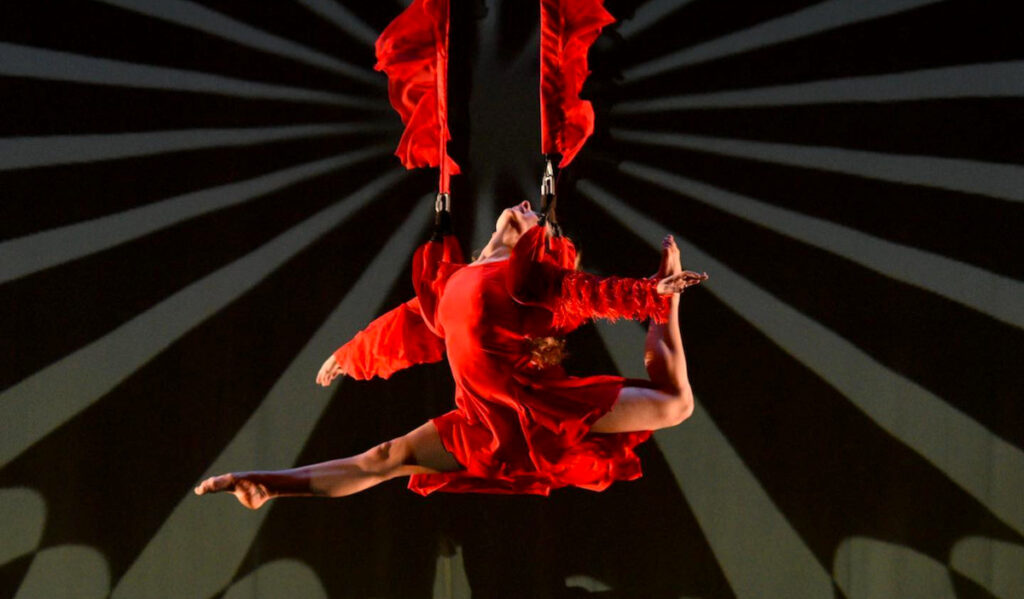 A dancer in red suspends from the ceiling on two ropes with a sunburst pattern behind her