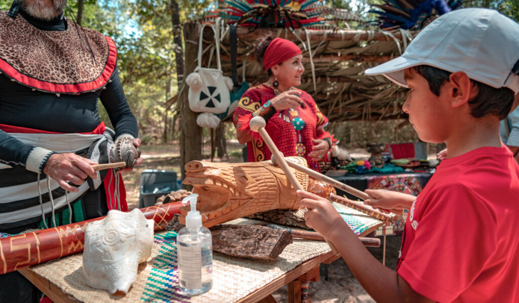 A child plays an Indigenous instrument with costumed demonstrators