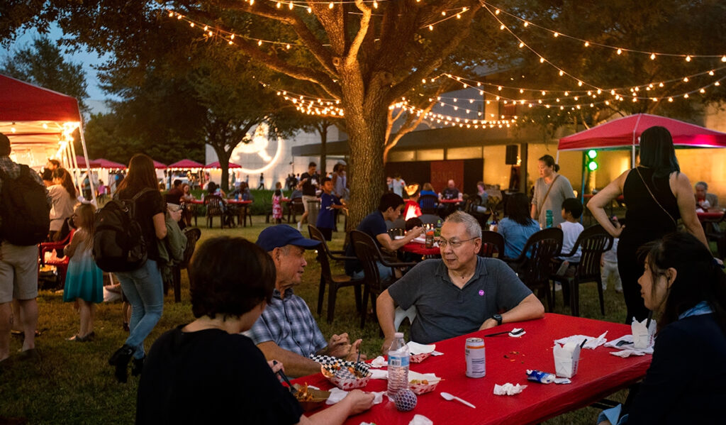 People sit around tables under trees and strings of lights