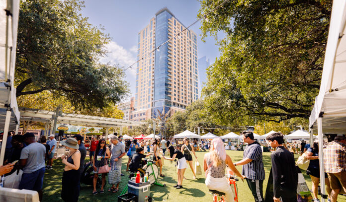 An outdoor market with booths and people moving around under a tall skyscraper