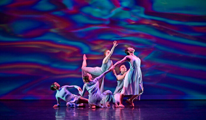 Five dancers cling to one another in front of a swirling, colorful backdrop