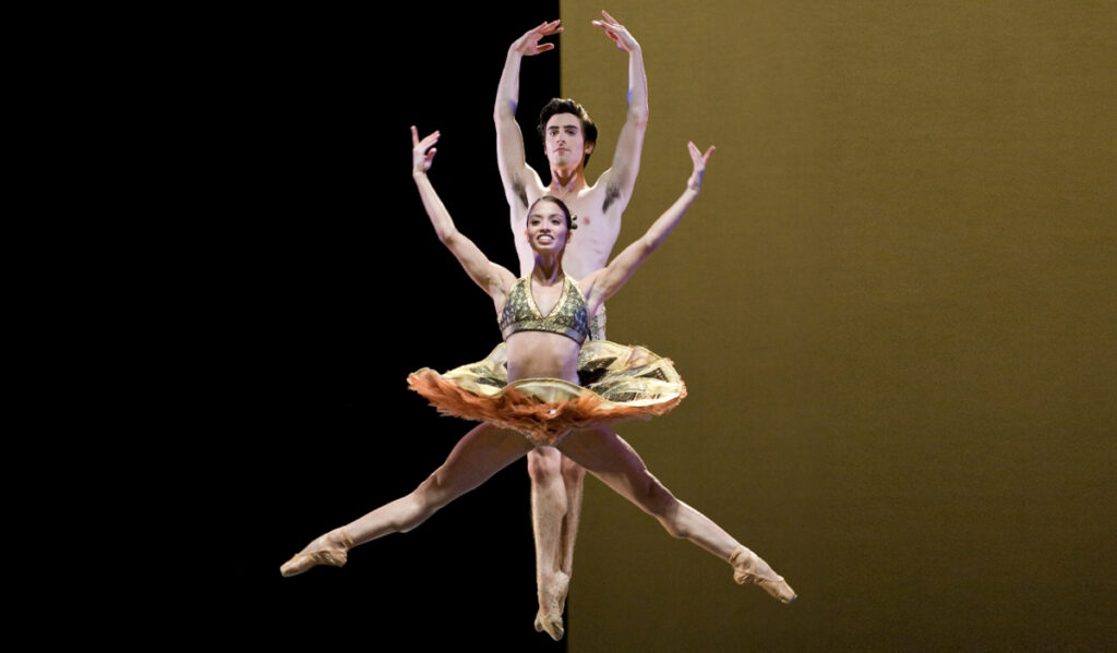 A ballerina leaping with arms and legs spread as a male ballerina leaps behind her