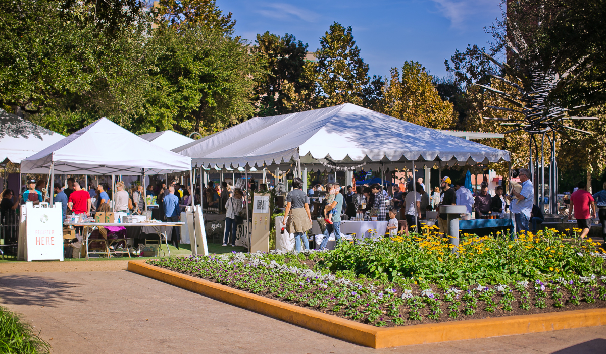 A market scene on a sunny day with people walking by landscaped gardens and booths