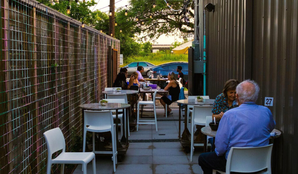 A narrow patio with small tables and chairs and scattered groups of people
