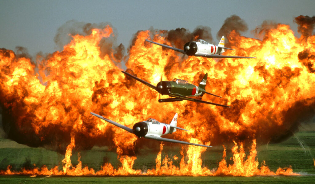 Three World War II planes fly past a wall of flames